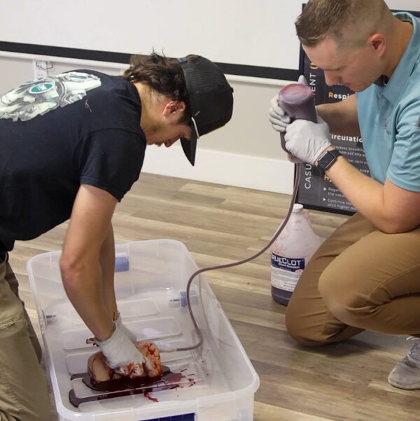 Two individuals participate in a hands-on medical training exercise, simulating wound packing with artificial blood and a realistic leg wound model in a classroom setting.