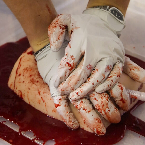 Close-up of gloved hands applying pressure to a bleeding wound simulator during a medical training exercise.